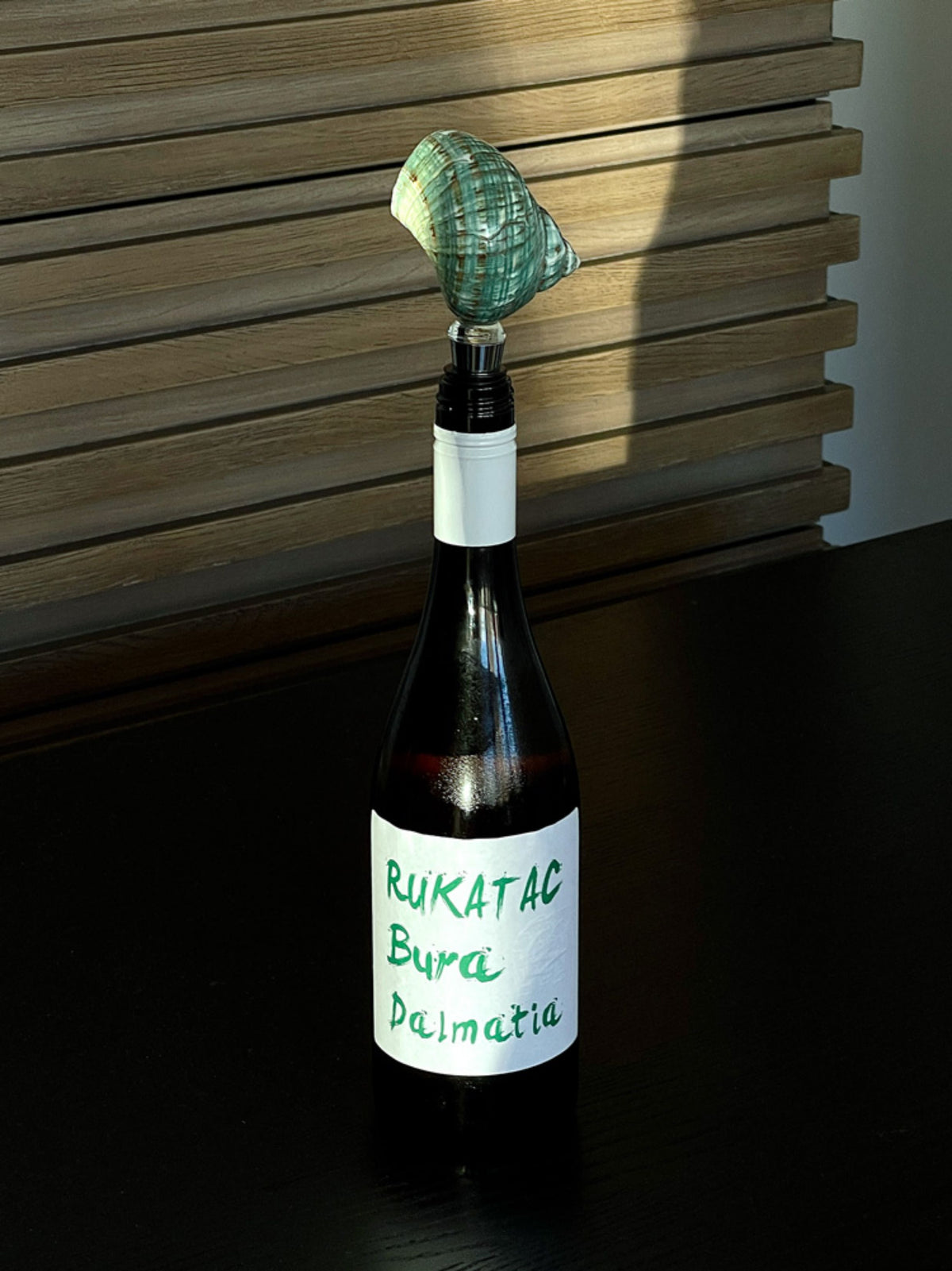 Green spiral seashell wine stopper in a dark wine bottle by wooden blinds