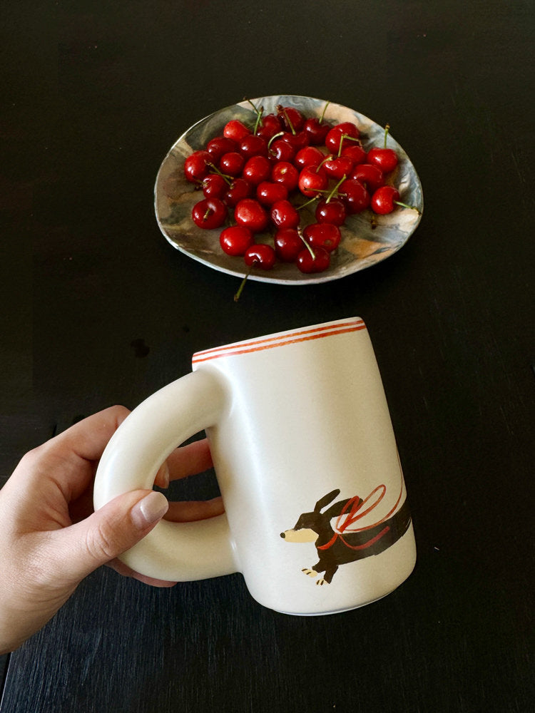 Hand holding a SoftServe dachshund ceramic mug above a table with cherries